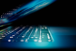 Ein halb zugeklappter Laptop in dunklem Hintergrund mit sich anleuchtenden Bildschirm und Tastatur